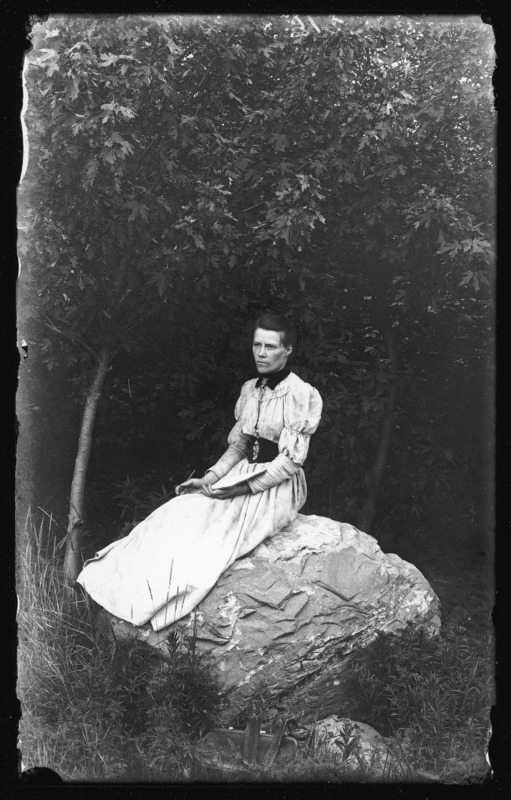 A woman, Lucy Pascal, holding a sketch pad. Lucy is the sister of Descartes Pascal. She is sitting on a boulder under a tree.