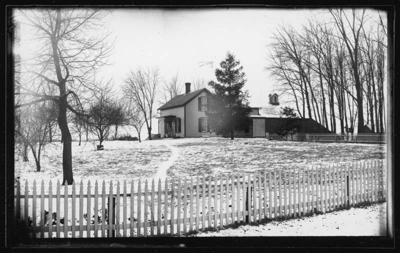 The Bussen farm house and outbuildings in winter. The house and yard are enclosed by a picket fence. The lawn is covered with snow. There is cupola on one of the outbuildings.