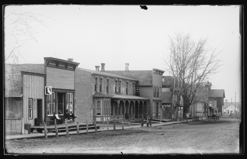 Street scene in Grand Mound, Iowa. Two men are sitting outside a storefront. Above another shop there is a sign for a cobbler named "A. Schultz" and a dog is sitting beside the doorway to the shop. Farther down the street, three young boys lean against a fireplug or hydrant.