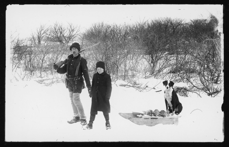 Hunter with boy and dog standing outside a thicket. On the left is Wickliffe Cotton, holding a rifle and wearing an ammunition belt. Wickliffe was the cousin of Descartes Pascal and the son of Salem and Mercy Cotton. On the right is Lee Pascal, nephew of Descartes Pascal. A dog named Pedro sits on a handmade sled beside three dead rabbits. Snow covers the ground.