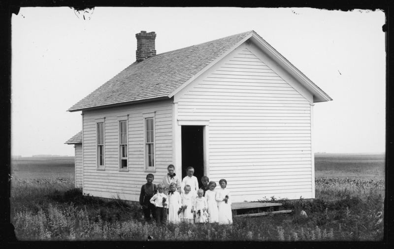 Teacher and students posed outside a one-room schoolhouse. The children in the front row are dressed in white and are carrying flowers. The tallest woman on the left in the back row is Lucy Pascal, sister of Descartes Pascal. The school is located in Lincoln Township, Pocahontas County, Iowa.