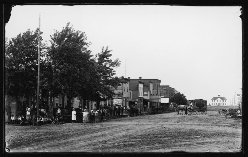West side of the main street in Pocahontas, Iowa. There are many men, women, children, wagons, and horses in this street scene.
