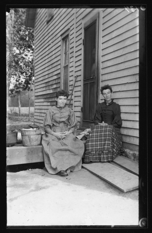 Two women seated beside the back door of their house in DeWitt, Iowa. On the left is Della M. Cotton and on the right is Bertha M. Cotton. They are the daughters of Salem Cotton and his wife Mercy Brown.