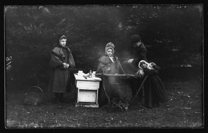 A boy pours something from a bucket into a cauldron steaming over a fire. The girls are dressed in coats and bonnets.