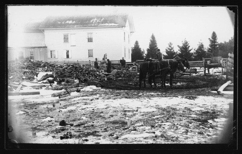 Back view of the Pascal house, Grand Mound, Iowa in winter. Men are cutting wood. Between the two men on the left is a circular saw. On the right are two horses.
