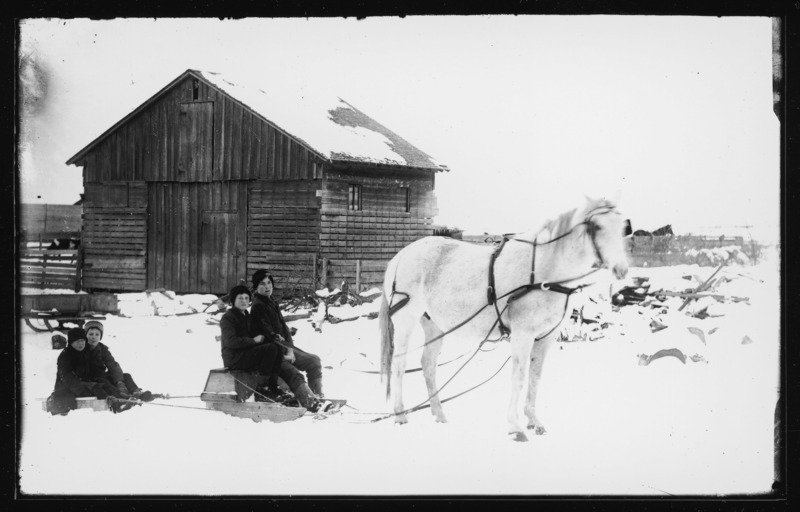 Boys on horse-drawn sleds in winter. Lee Pascal and Jasper Babcock are on the front sled. Percy Pascal and Jim Townsen are on the rear sled. The horse's name is Daisy. The photo is taken in front of the corn crib on the Pascal farm.