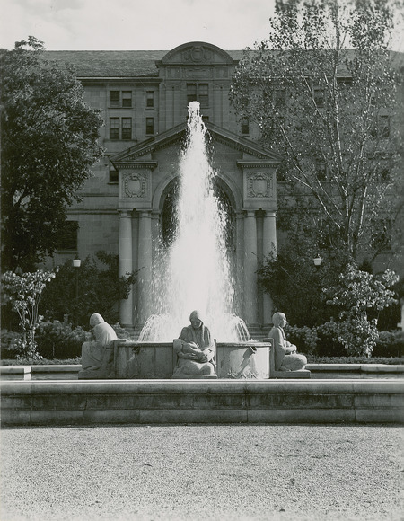 "Four Seasons" fountain with the Memorial Union in the background. The figure facing front nurses her newborn, the figure facing right holds a basket of maize, and the figure facing left plants the seed corn. The fourth figure (not visible) holds the tender shoot. Cco: Silver gelatin photograph on board