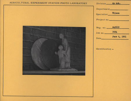 Photograph of the round plaster positive and negative plaque sitting on a shelf. The plaster has a center image of a woman who wearing a long dress, with smaller images along the edge and across the middle of the plaque. In front of them there is a clay figure of a mother and son.