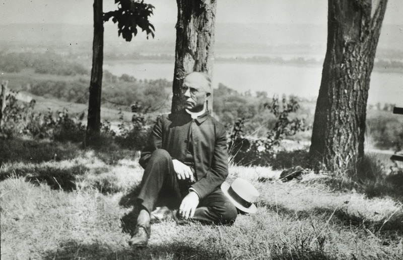 Dr. Leroy Titus Weeks, who appears to be wearing clergy attire, sits on the ground leaning against a tree. Slide originally titled "Dr. Leroy Titus Weeks."