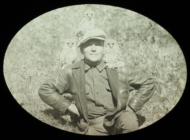 Walter Rosene sitting on the ground, posing with three owls, April 28, 1929. Rosene has an owl perched on each of his shoulders and one owl perched on the top of his head, sitting on his hat. Slide originally titled "Rosene with Owls."