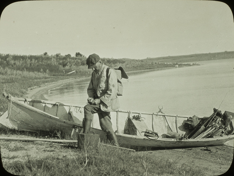 Walter Rosene standing next to a boat at Stump Lake, North Dakota. Slide originally titled "W.M. Rosene at boat Stump Lake, N.D." This item is related to Rosene's journal: https://n2t.net/ark:/87292/w9jd4pp05.