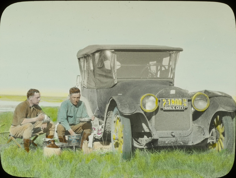 Walter Rosene and Walter W. Bennett sitting next a car eating lunch. Slide originally titled "Walter Rosene and W. Bennett--Lunch." The lantern slide is hand-colored.