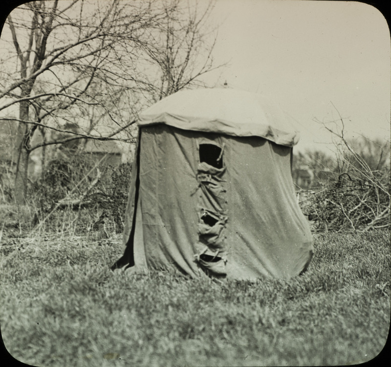 An umbrella blind used for photographing birds, set up in a grassy area. The blind has an entrance on the left side and several different apertures through which photographs can be taken. Slide originally titled "Blind for Photographing Birds." Details provided on the back of photograph.