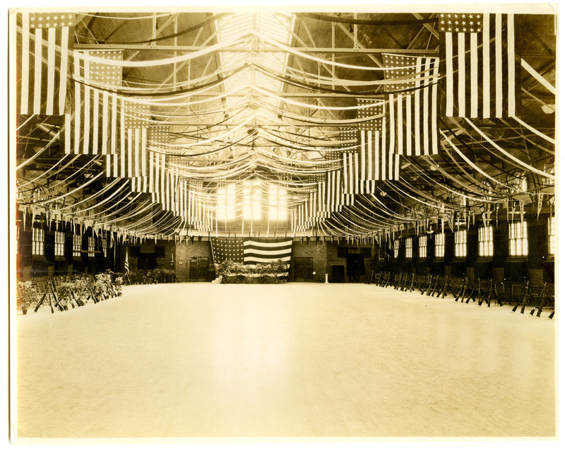 State Gymnasium is decorated for a military ball. One large American flag is hanging down from the second floor. Other flags and streamers are hanging from the ceiling. Rifles are stacked in a tripod formation along the sides of the gymnasium.