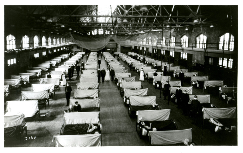 Rows of cots line the State Gymnasium floor to accommodate soldiers suffering from the Spanish influenza epidemic. The gym is full and some of the healthy soldiers are assisting the sick ones.