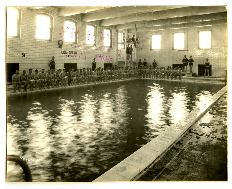 Men are sitting along the edge of the swimming pool in State Gymnasium. Other men are standing behind them or on the diving boards.