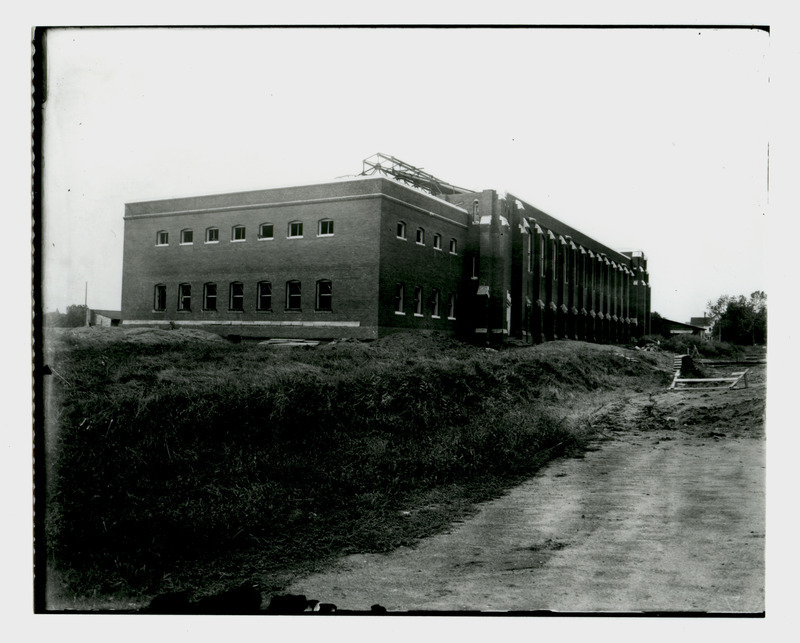 At this stage of construction the exterior brick walls and roof trusses of State Gymnasium are visible in this northeast view. Piles of dirt and building supplies surround the gymnasium. There are no sidewalks or roads.