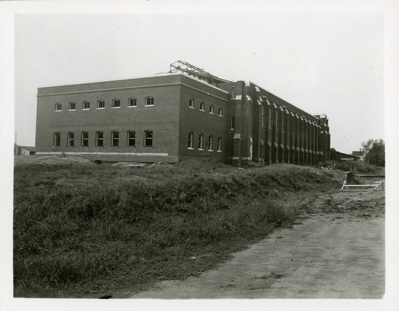 At this stage of construction the exterior brick walls and roof trusses of State Gymnasium are visible in this northeast view. Piles of dirt and building supplies surround the gymnasium. There are no sidewalks or roads.