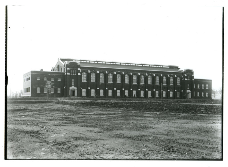 Two men are working in the middle of the first floor windows on the front (north) side of State Gymnasium. The left doorway is partially open. A dirt road runs along the front of the building.