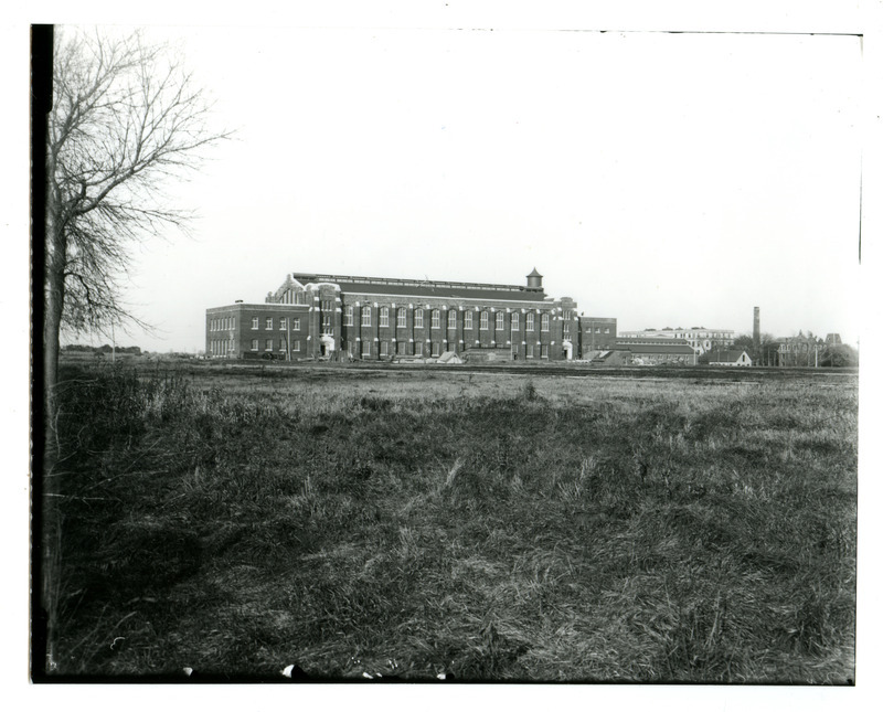 Viewed from a distance, the back (south) side of State Gymnasium is visible. Lab of Mechanics, Machine Shop, Quarantine Hospital and Main Building (Beardshear Hall) can be seen on the far right. The Marston Water Tower is in the background. Building supplies are laying around the building.