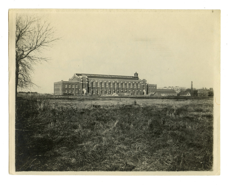 Viewed from a distance, the back (south) side of State Gymnasium is visible. Lab of Mechanics, Machine Shop, Quarantine Hospital and Main Building (Beardshear Hall) can be seen on the far right. The Marston Water Tower is in the background. Building supplies are laying around the building.