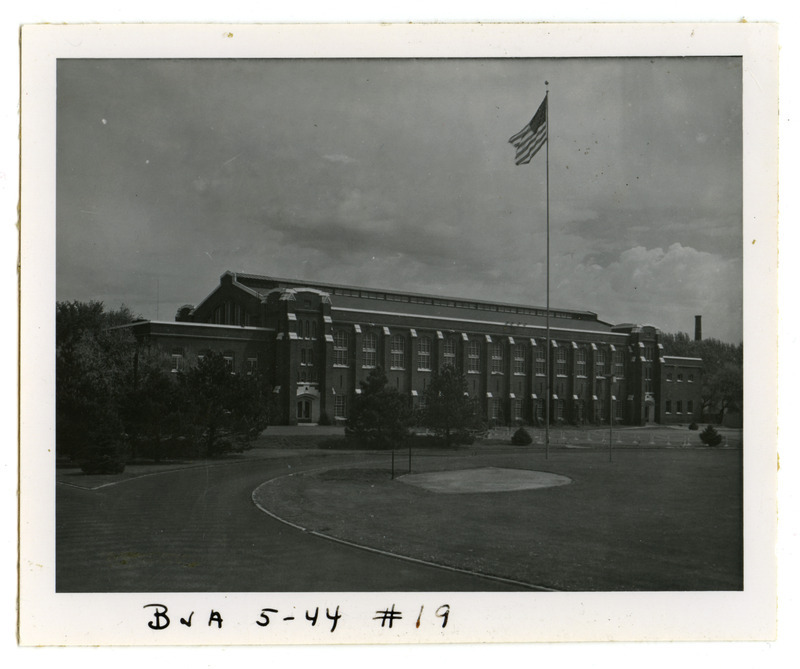 The track surrounds State Field (Clyde William Field) in this southwest view of the back (south) side of State Gymnasium. Hurdles are positioned on the other side of the track. A sand pit, possibly used for a track and field event, is located in front of the flag pole.