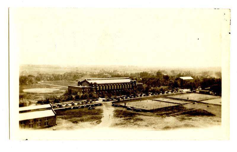 This aerial view of the State Gymnasium is labeled "in front of Mens Gymnasium, Iowa State College." Part of the track and stadium is visible in back, and athletic courts are in use by people across the road in front. Cars from the 1920s or 1930s line the road in front. The Machine Shop is visible on the left, and neighborhood buildings are visible across Sheldon Avenue on the right.