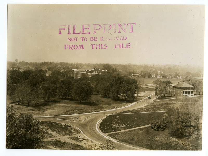 The State Gymnasium is viewed from the intersection of Welch Road and Union Drive, looking west, with the College Hospital on the right. A wagon with two horses is driving on the road that passes by a grove of trees where Friley Hall now stands. Surrounding trees and houses on the west side of campus are also visible.