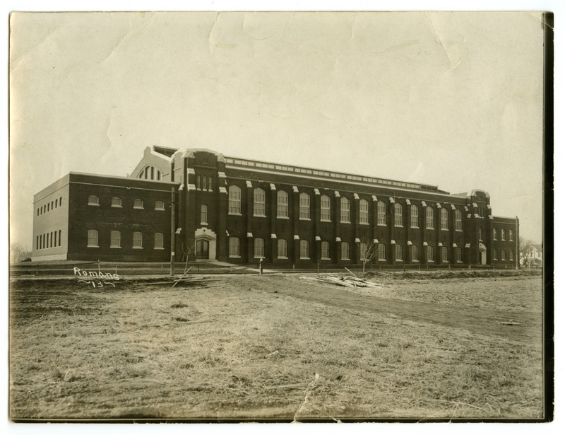 The front of the newly constructed State Gymnasium is viewed from the northeast, with two small leafless trees in front and a house on the right.