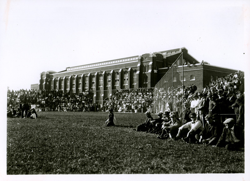 Spectators fill bleachers behind the chain link fence backstop of the new State Field (later Clyde Williams Field). The State Gymnasium is in the background with some spectators sitting on the roof of the east wing. Others overflow from the bleachers and sit on the field.