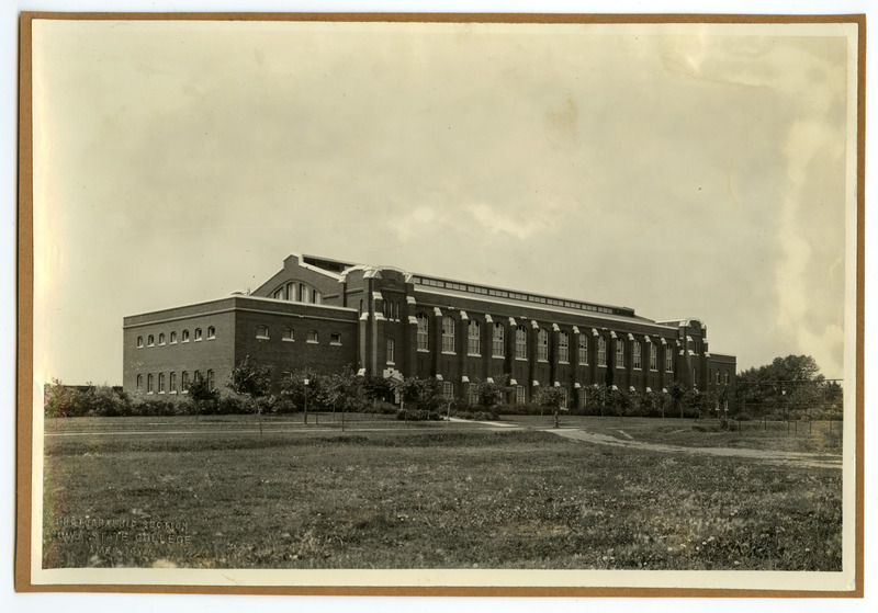 The front of the State Gymnasium is viewed from northeast with small trees growing along the road in front and the fence of an athletic field on the right.