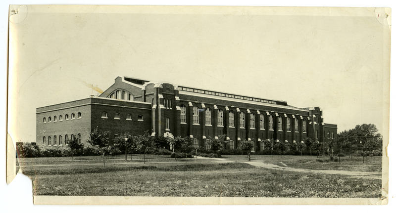 The front of the State Gymnasium is viewed from northeast with small trees growing along the road in front and the fence of an athletic field on the right.