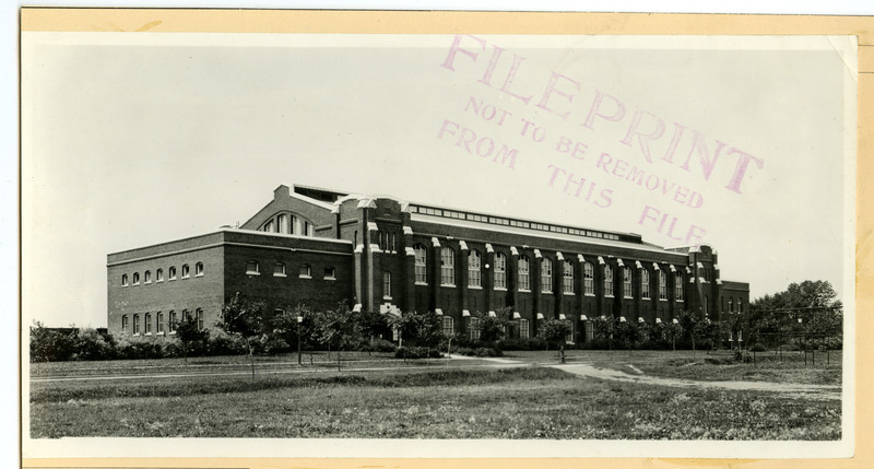 The front of the State Gymnasium is viewed from northeast with small trees growing along the road in front and the fence of an athletic field on the right.