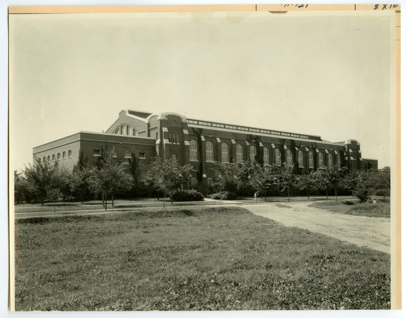 The front of the State Gymnasium is viewed from northeast with trees hiding part of the building. The fence of an athletic field is on the right with people sitting in the corner, and a dirt road leads past it towards the building.