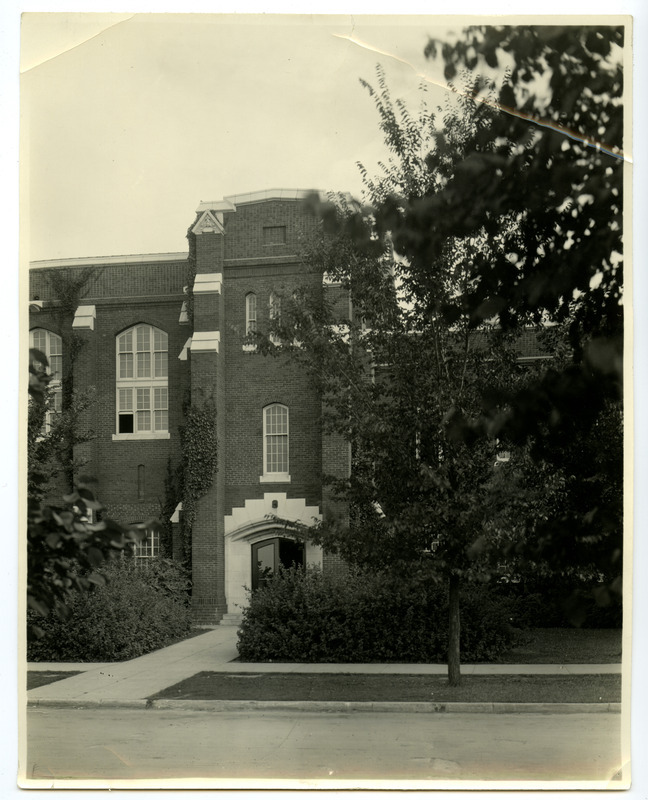 The front (north) entrance on the west end of the State Gymnasium is marked by a tower, with an open door on the right and an open window on the upper left. Trees partially obscure the building, and ivy climbs the left side of the tower in this darkened photograph.