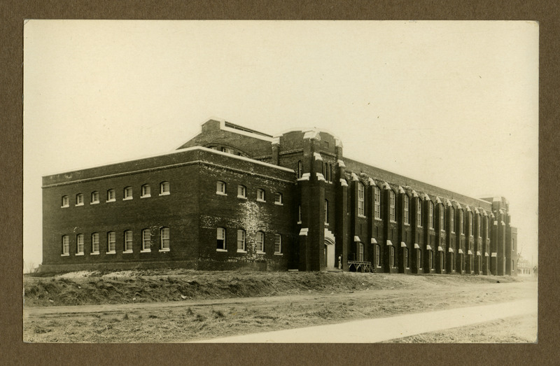 The State Gymnasium under construction is viewed from the northeast side of the front. A man stands by scaffolding near the northeast entrance, dirt surrounds the building, and some bricks show white efflorescence.