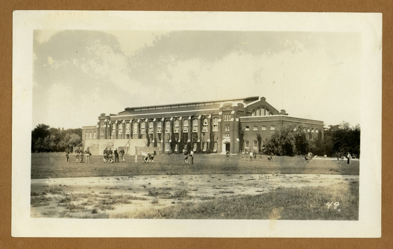 Football players practice in back of the State Gymnasium, viewed from the southeast side of the State Field (later Clyde Williams Field). The uniforms are from an early time period, and ivy grows up the sides of the building to the roof. Handball courts are also in back of the building.