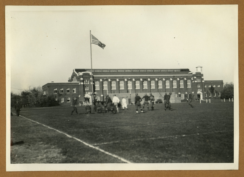 Football players practice in back of the State Gymnasium, viewed from the southwest side of the State Field (later Clyde Williams Field). The uniforms are from an early time period. A 1920s car is parked near the handball courts, and the flag waving over the field has 48 stars.