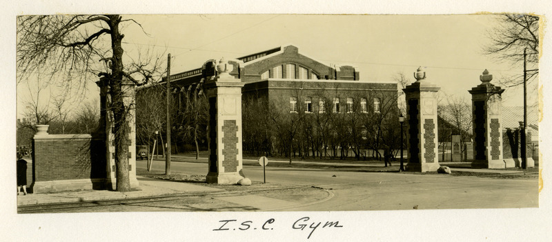 The State Gymnasium is viewed through the West Gate brick pillars, with a 1920s car parked in front of the building.