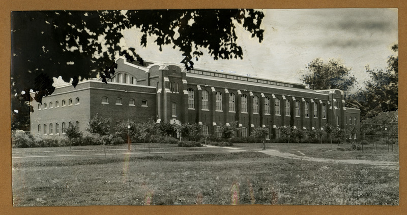 The front of the State Gymnasium is viewed from northeast with small trees growing along the road in front and the fence of an athletic field on the right, identical to photos taken in 1919. Trees have been painted in on the upper left corner and right side.