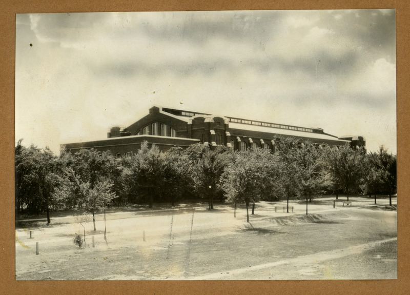 The front of the State Gymnasium is viewed from the northeast, showing medium-sized trees and new landscaping across the street to the north.