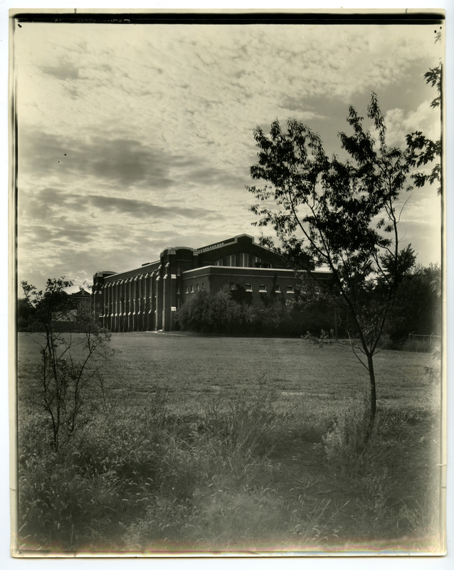 The back (south) side of State Gymnasium is viewed from the east with a small tree and brush in the foreground.