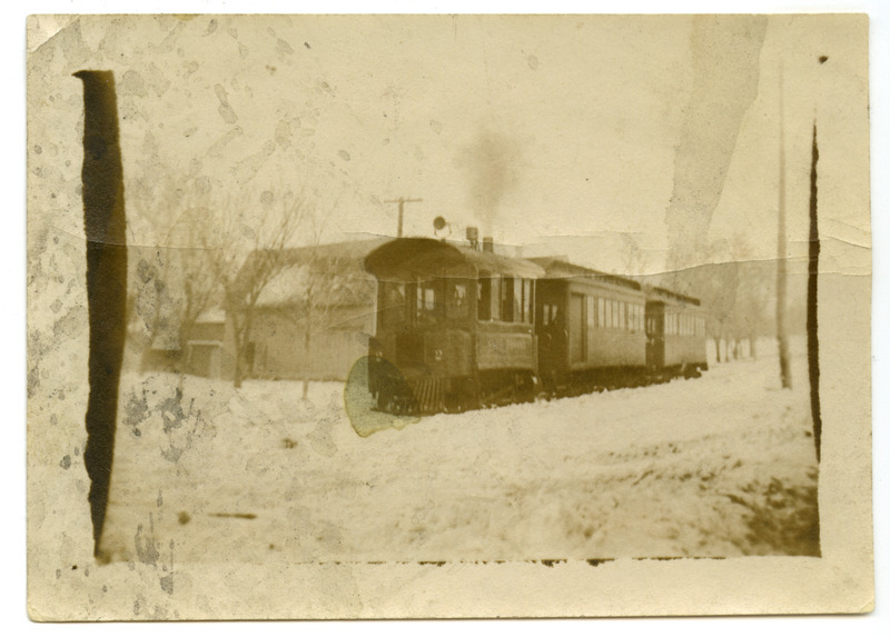 The Ames & College Railway (The Dinkey) locomotive and two cards are moving past several frame buildings.