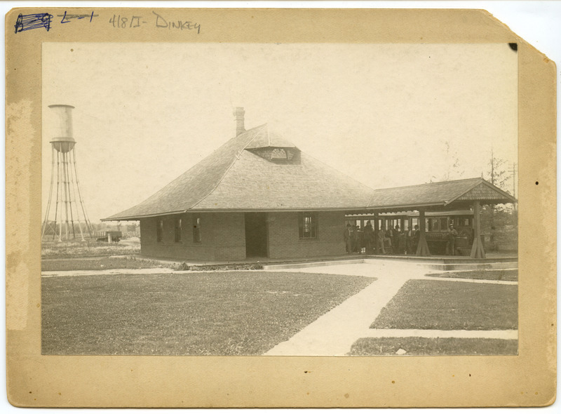 A group of passengers are standing on the station platform in front of the Ames & College Railway (The Dinkey). The Marston Water Tower is in the background.