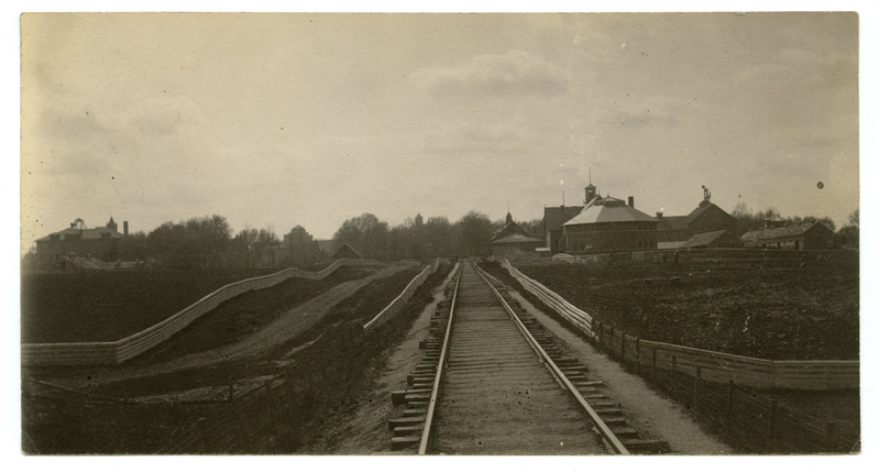 The Ames & Campus Railway (The Dinkey) tracks are running through campus. Various buildings and fences areas are on both sides of the tracks. One fences area contains a herd of small grazing animals, perhaps goats or sheep. Two individuals are also in this fenced area.