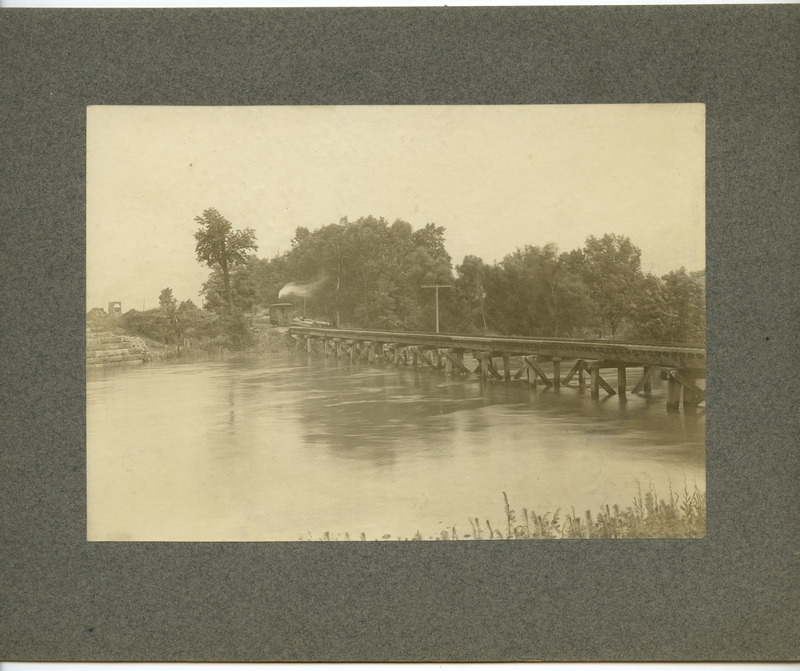 The Ames & College Railway (The Dinkey) engine is attempting to cross a over Squaw Creek. The creek is high, but not covering the bridge. The Chicago & Northwestern (C&NW) coal chute is in the distance.