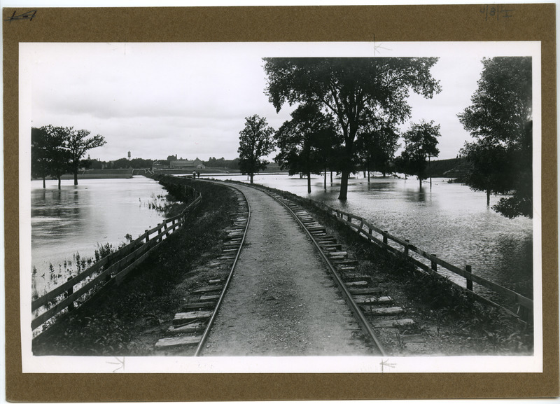 During a flood occuring in Spring 1901 or 1902, water in Squaw Creek over-flowed the banks, submerging the surrounding area. The water is high, but doesn't appear to be over the Ames & college Railway (The Dinkey) tracks. Two individuals are observing the creek and flood. The Marston Water Tower, Carrie Chapman Catt Hall (Botany Hall) and Old Main (after the 1st fire) are in the distance.