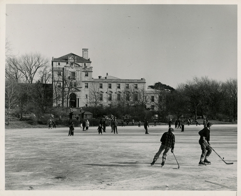 Two men playing ice hockey on a frozen Lake LaVerne. People are skating in the background. Farther back the Memorial Union is seen.