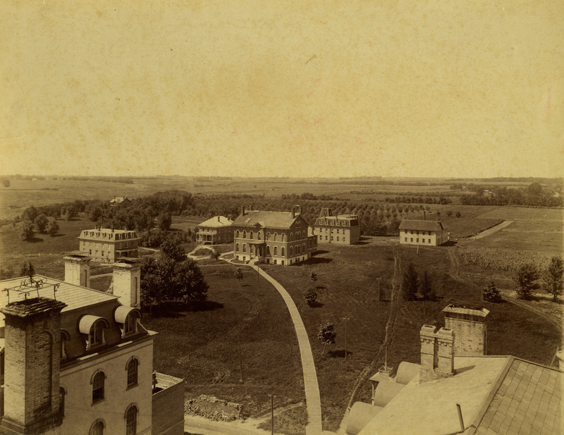 View southwest from Main Building. The sidewalk leads to Chemical and Physical Laboratory, built in 1870 & 75, it burned in 1913. East and West Cottages, men's dormitories, are to the left of it. East Cottage, built in 1882, was later used as Training Quarters for athletes. It was torn down in 1907. West Cottage, built in 1880, was later used by the College hospital. It was torn down in 1934. In back of Chemical Hall is the Engineering Building, built 1883 & 1884. Later it was known as the Theoretical and Applied Mechanics Laboratory and now known as Mechanics Laboratory. To the right of Engineering Hall is the frame workshop, built in 1870. It was torn down in 1897. The house in the trees is the Marston Cottage which was torn down after Mrs. Marston's death in 1958 when it was almost surrounded by Friley Hall.Tennis courts and the football field supplanted the orchards.