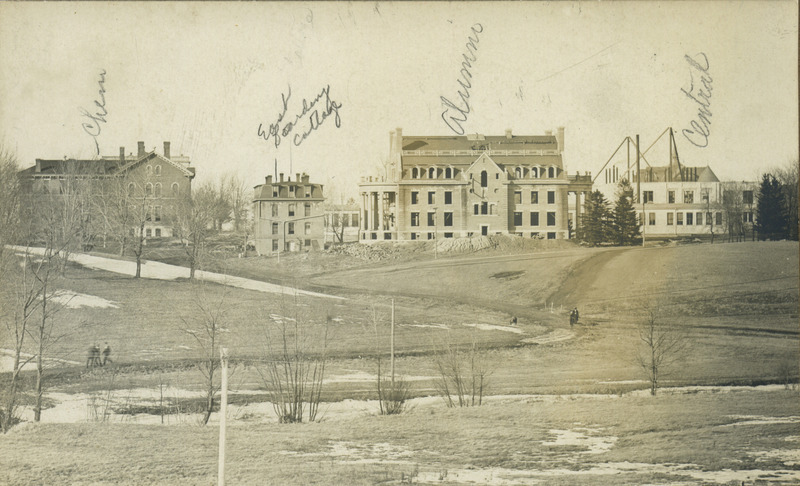 Campus buildings viewed from left to right: Chemical and Physical Laboratory; East Boarding Cottage, Alumni Hall, and Central Building (later known as Beardshear Hall). Alumni Hall and Central Building are under construction. There are people walking along the dirt paths, 1906.